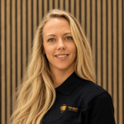 Portrait of a smiling woman with long blonde hair, wearing a black polo with TEMBO Safari logo, standing in front of a wooden slat backdrop.